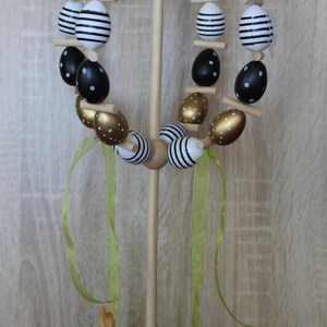 May include: A wooden Easter egg wreath featuring black, gold, and white painted eggs. The eggs are decorated with stripes and polka dots. The wreath is on a wooden stand with a small bunny figurine and a carrot at the base. Green ribbons are attached.