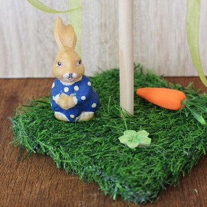 May include: An Easter decoration featuring a tan bunny in a blue polka-dot outfit on a patch of artificial grass. The scene includes a wooden post, an orange carrot, and a green flower, evoking a springtime theme.