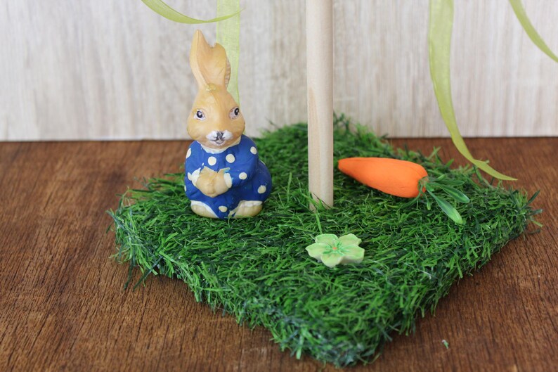 May include: An Easter decoration featuring a tan bunny in a blue polka-dot outfit on a patch of artificial grass. The scene includes a wooden post, an orange carrot, and a green flower, evoking a springtime theme.