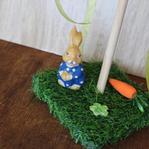 May include: A ceramic Easter bunny figurine in a blue polka-dot outfit, on a patch of green artificial grass. An orange carrot and a small green flower are also on the grass. A wooden dowel and green ribbon are in the background.