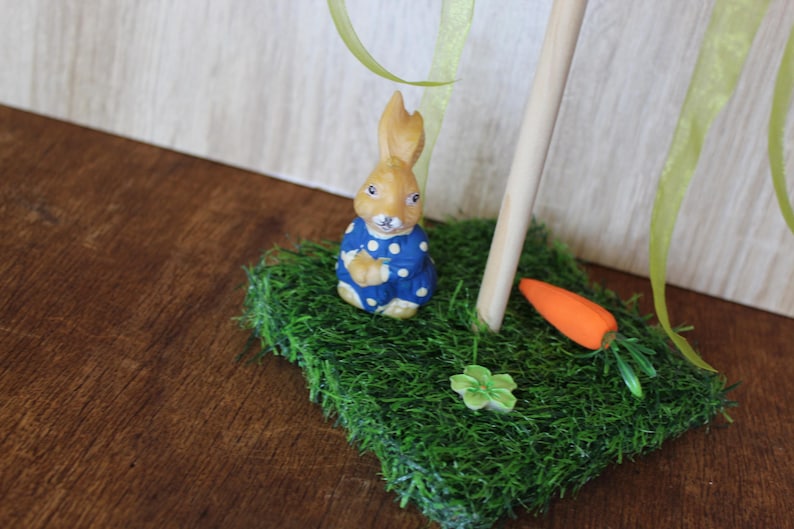 May include: A ceramic Easter bunny figurine in a blue polka-dot outfit, on a patch of green artificial grass. An orange carrot and a small green flower are also on the grass. A wooden dowel and green ribbon are in the background.