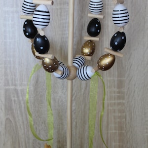 May include: A wooden Easter egg display with a circular arrangement of decorative eggs. The eggs are painted with gold, black and white patterns. A small bunny figurine and carrot are at the base, with green ribbons and a butterfly on top.