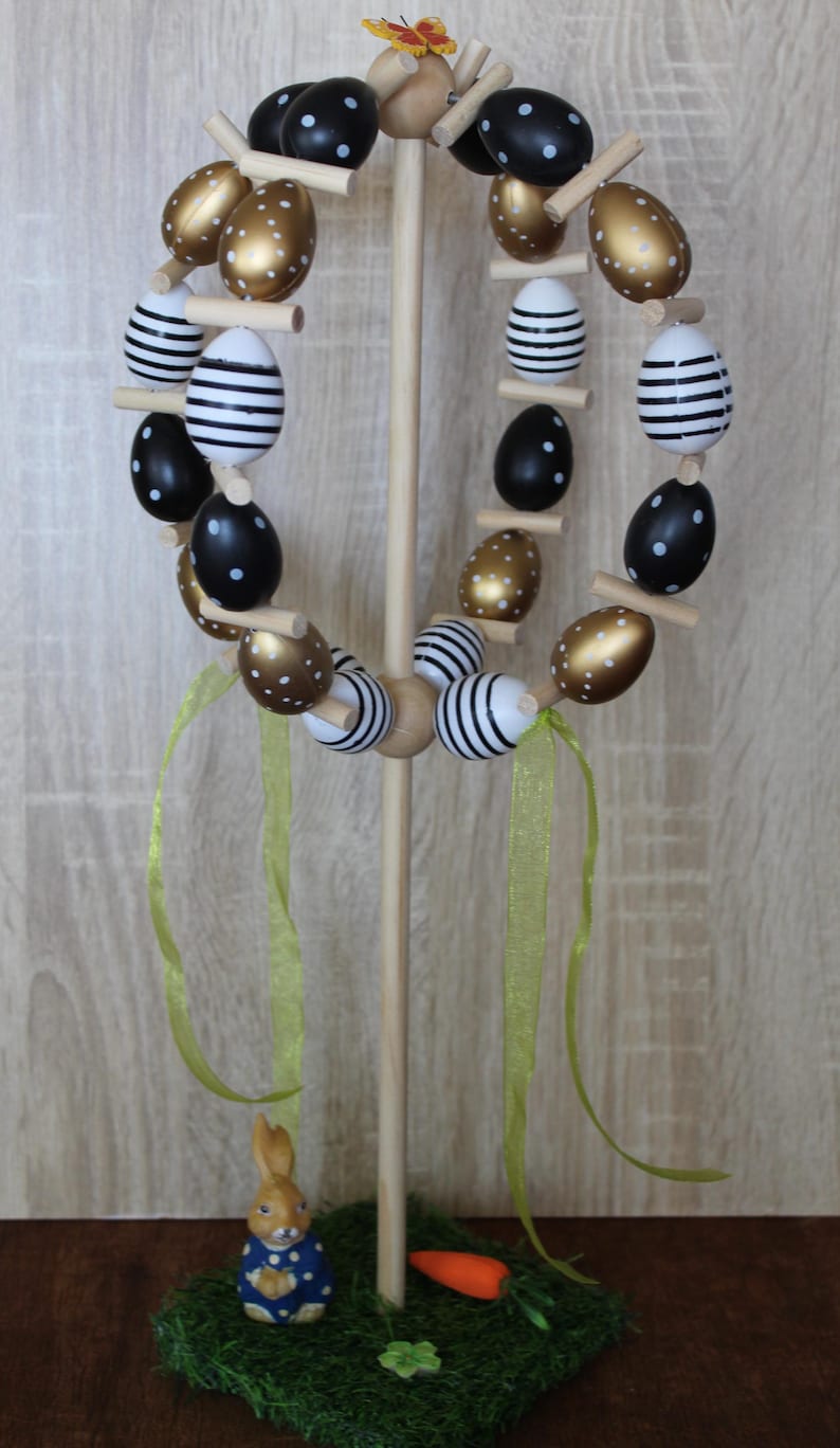 May include: A wooden Easter egg display with a circular arrangement of decorative eggs. The eggs are painted with gold, black and white patterns. A small bunny figurine and carrot are at the base, with green ribbons and a butterfly on top.