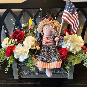May include: A patriotic centerpiece featuring a doll holding an American flag, surrounded by red and white flowers, greenery, and a candle. The doll wears a dress with a star and stripe pattern, evoking a festive, Americana theme.