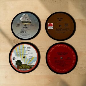 May include: Four circular coasters made from vintage vinyl records. Each coaster features a different record label and album title, including "Outlaws," "Free," "Traffic," and "Stephen Stills." The coasters have black edges and are arranged on a light wood surface.