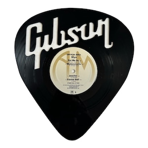 May include: A black Gibson guitar pick with a white Gibson logo. The pick is made from a vinyl record with a label that reads "Crystal Ball" by Styx, "Put Me On" by Jennifer, and "Mademoiselle" by Jennifer.
