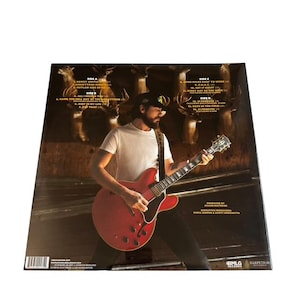 May include: A vinyl record album featuring a man playing a red electric guitar. The album cover has a background of deer and text listing song titles. The man is wearing a white t-shirt and a black cap.