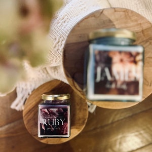 Könnte beinhalten: Zwei gläserne Kerzenbehälter mit goldenen Deckeln. Die Kerze links ist mit "RUBY" und einem floralen Design in Rosa- und Lilatönen beschriftet. Die Kerze rechts ist mit "JASMIN" und einem floralen Design in Blau- und Weißtönen beschriftet.