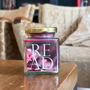 Könnte beinhalten: Ein Glas-Kerzengefäß mit goldenem Deckel und einem rosafarbenen Blumendesign. Das Gefäß trägt den Schriftzug "READ" in weißen Buchstaben. Die Kerze steht auf einem Holztisch.