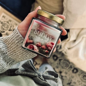 Könnte beinhalten: Eine Kerze in einem Glasgefäß mit goldenem Deckel. Das Etikett lautet "Bookalicious Candles" und "Buddy Bread" mit einem floralen Design.