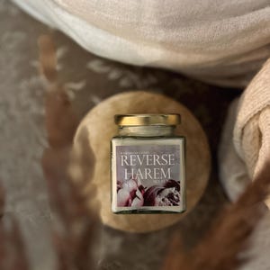 Könnte beinhalten: Ein Glas-Kerzengefäß mit einem weißen Etikett, auf dem in schwarzer Schrift "Reverse Harem" steht. Das Etikett zeigt auch ein Blumenmuster mit rosa und violetten Blumen. Das Kerzengefäß steht auf einer Holzoberfläche.