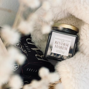 Könnte beinhalten: Ein Glas-Kerzengefäß mit goldenem Deckel und einem weißen Etikett mit der Aufschrift "Bookalicious Candles Reverse Harem Wood Sage & Sea Salt Type". Die Kerze steht auf einem weißen Kunstfell.