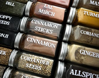 Könnte beinhalten: Nahaufnahme verschiedener Gewürzgläser mit silbernen Deckeln. Die Gläser sind mit weißem Text beschriftet, darunter "Bay Leaves", "Oregano", "Dill", "Poppy Seeds", "Cinnamon Sticks", "Cinnamon", "Coriander Seeds" und "Ground Cumin". Die Gewürze sind sichtbar.