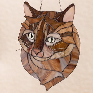 May include: A stained glass window hanging with a chain, featuring a close-up of a brown tabby cat's face. The cat's eyes are green, and the glass is cut into small pieces to create a mosaic effect.