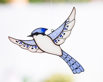May include: A stained glass blue jay bird ornament hanging from a suction cup on a window. The bird is in flight with its wings spread and has blue, white, and black details.