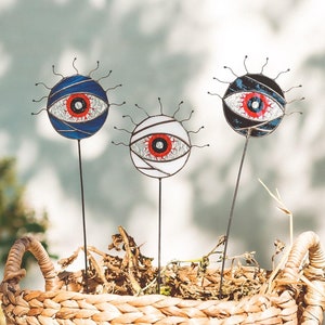 May include: Three decorative eye-shaped garden stakes with blue, white, and black glass accents. The stakes are placed in a woven basket filled with dried foliage.