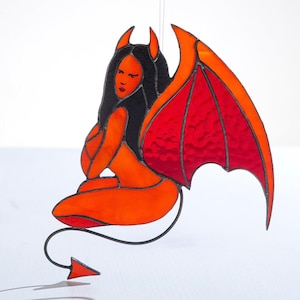 May include: A stained glass depiction of a red devil with wings and a tail. The devil is sitting with its legs crossed and has a mischievous expression on its face.