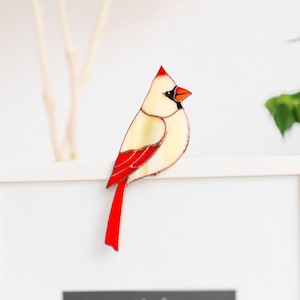 May include: A stained glass cardinal ornament featuring a cream-coloured body, a bright red wing, and a long red tail. The bird has a black face mask and a red beak, perched on the top edge of a white frame containing the text sometimes you win / sometimes