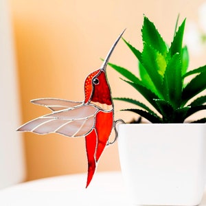 Puede incluir: Un colibrí de vidrio emplomado con alas rojas y blancas, posado en una maceta blanca con una planta suculenta verde.