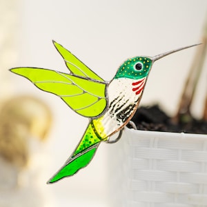 Pode incluir: Um beija-flor de vidro colorido com asas verdes, amarelas e brancas. O beija-flor está empoleirado em um vaso branco com um padrão tecido.