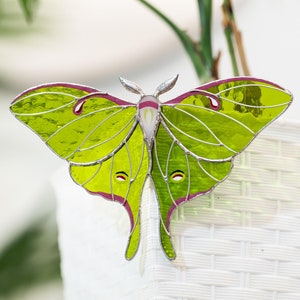 May include: A green and purple stained glass luna moth with a white body. The moth is sitting on a white wicker planter.