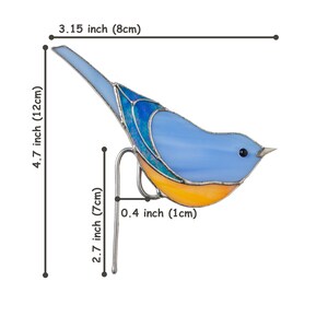 以下が含まれることがあります： オレンジ色のアクセントが付いた青いステンドグラスの鳥。高さ12cm、幅8cm。鳥は高さ7cmの金属製のスタンドに取り付けられています。