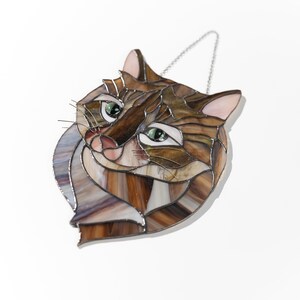 May include: A stained glass cat head with brown, tan, and white glass pieces. The cat has green eyes and a pink nose. The cat is hanging from a silver chain.