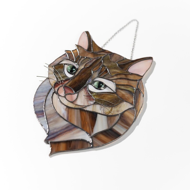 May include: A stained glass cat head with brown, tan, and white glass pieces. The cat has green eyes and a pink nose. The cat is hanging from a silver chain.