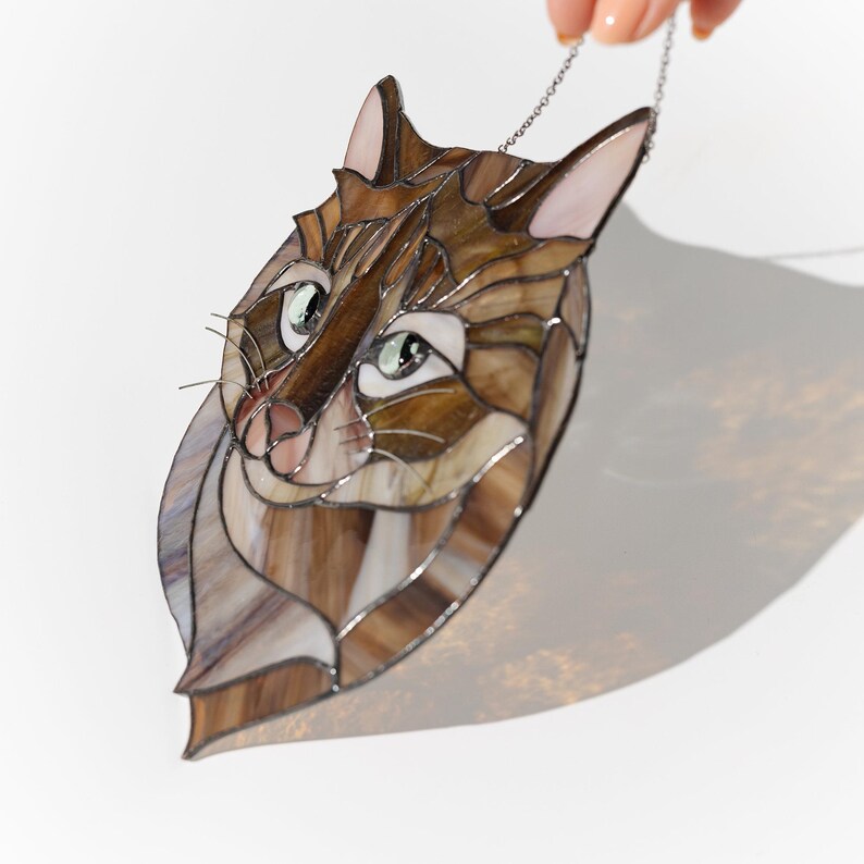 May include: A stained glass cat head with brown, white, and black colors. The cat has green eyes and a pink nose. The cat head is hanging from a silver chain.