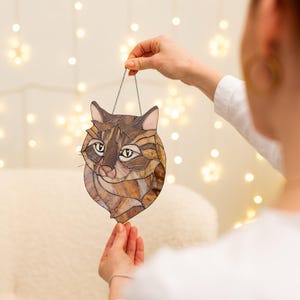 May include: A stained glass cat head suncatcher with brown, black, and white glass pieces. The cat has yellow eyes and a pink nose.