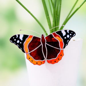 Könnte beinhalten: Ein Buntglas-Schmetterling mit detailliertem Design. Der Schmetterling hat einen dunkelroten Körper, orange und schwarze Flügel mit weißen Punkten und silberfarbenen Drahtakzenten. Der Schmetterling wird vor einem verschwommenen grünen Hintergrund präsentiert.