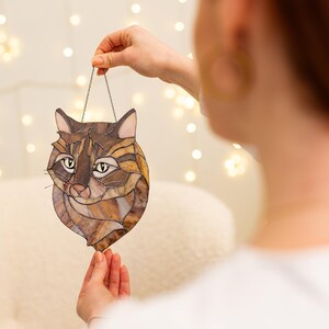 May include: A stained glass cat head hanging from a silver chain. The cat has brown, black, and white fur and yellow eyes.