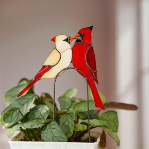 May include: A stained glass ornament featuring two cardinal birds perched on a metal arch. One bird is cream-colored with red wing accents, while the other is bright red. Both birds have black facial details and orange beaks, joined by dark metal solder lines.