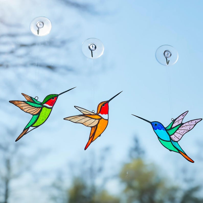 K&ouml;nnte beinhalten: Drei farbenfrohe Kolibri-Fensterbilder, die an Saugn&auml;pfen h&auml;ngen. Die Kolibris weisen leuchtende Farben auf: Gr&uuml;n, Rot, Orange, Blau und Lila. Jeder Vogel hat einen langen, schlanken Schnabel und ausgebreitete Fl&uuml;gel, was eine dekorative Fensterdekoration ergibt.