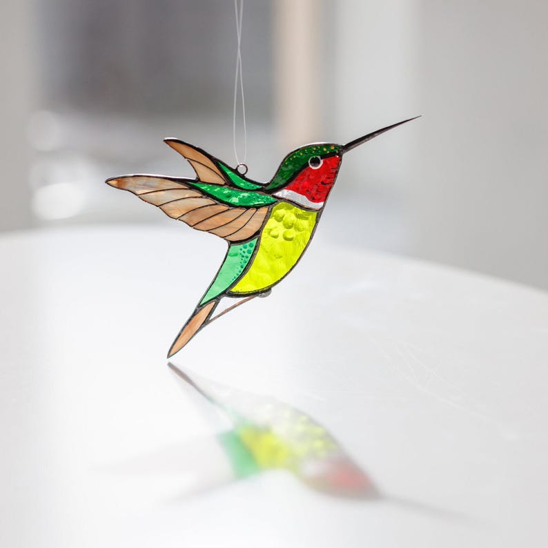 K&ouml;nnte beinhalten: Ein Glasmalerei-Hummel-Ornament mit rotem Hals, gr&uuml;nem Kopf und gelbem K&ouml;rper. Die Fl&uuml;gel sind braun und klar. Der Vogel h&auml;ngt an einem Faden und wirft einen farbenfrohen Schatten auf die Oberfl&auml;che.