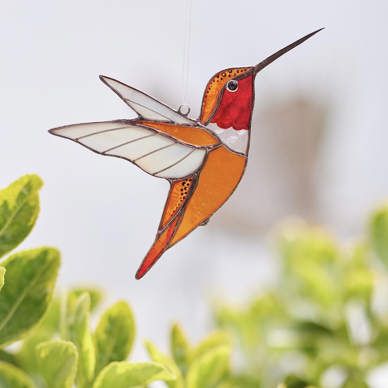 K&ouml;nnte beinhalten: Ein Kolibri-Anh&auml;nger aus Buntglas mit orangefarbenen, roten und wei&szlig;en Akzenten. Der Kolibri ist im Flug mit ausgebreiteten Fl&uuml;geln.