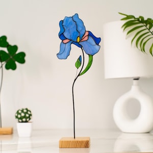May include: A stained glass blue iris flower sculpture on a wooden base. The flower is detailed with black lines and a pink center.