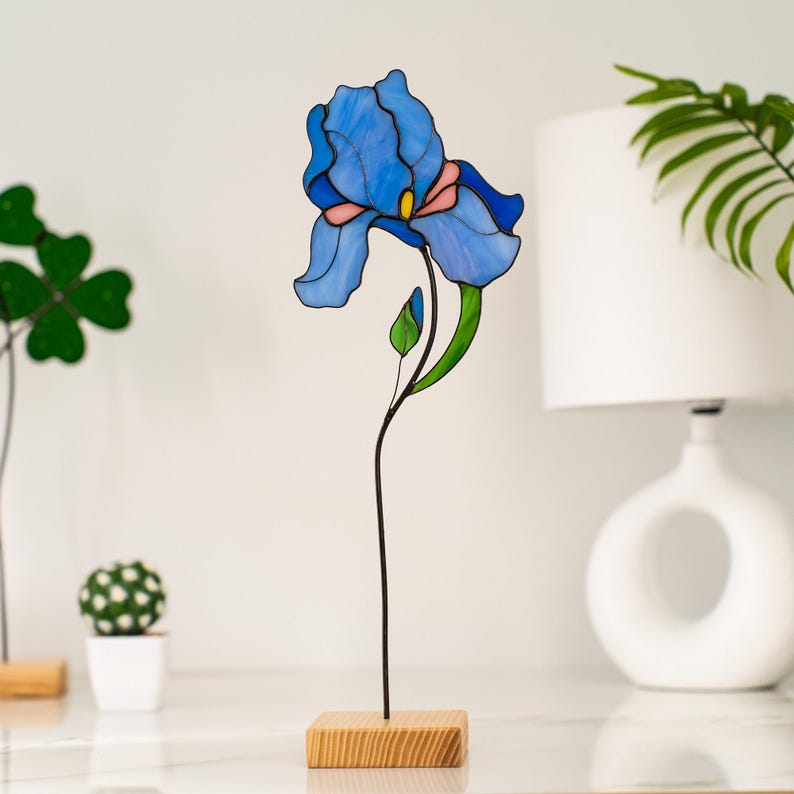 May include: A stained glass blue iris flower sculpture on a wooden base. The flower is detailed with black lines and a pink center.