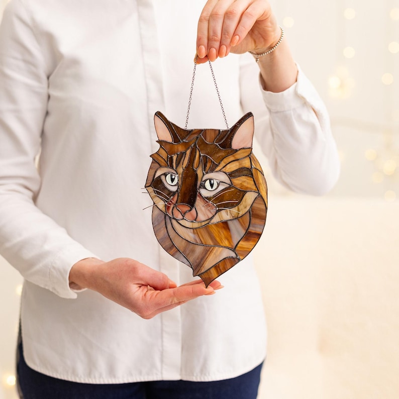 May include: A stained glass cat head with brown, orange, and white glass pieces. The cat has a light brown nose and green eyes.