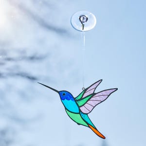 K&ouml;nnte beinhalten: Ein farbenfroher Kolibri-Suncatcher mit blauen, gr&uuml;nen und lila Akzenten. Der Vogel h&auml;ngt an einem Saugnapf, ideal f&uuml;r die Fensterdekoration. Der Kolibri hat einen langen, schwarzen Schnabel und ausgebreitete Fl&uuml;gel.