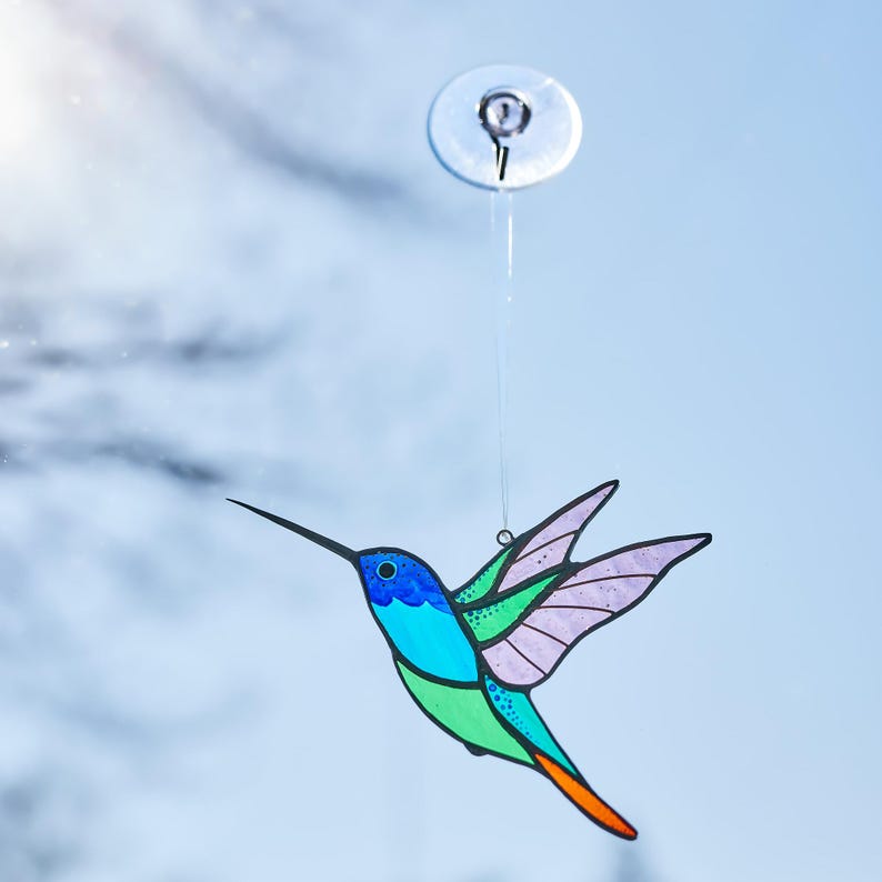 K&ouml;nnte beinhalten: Ein farbenfroher Kolibri-Suncatcher mit blauen, gr&uuml;nen und lila Akzenten. Der Vogel h&auml;ngt an einem Saugnapf, ideal f&uuml;r die Fensterdekoration. Der Kolibri hat einen langen, schwarzen Schnabel und ausgebreitete Fl&uuml;gel.