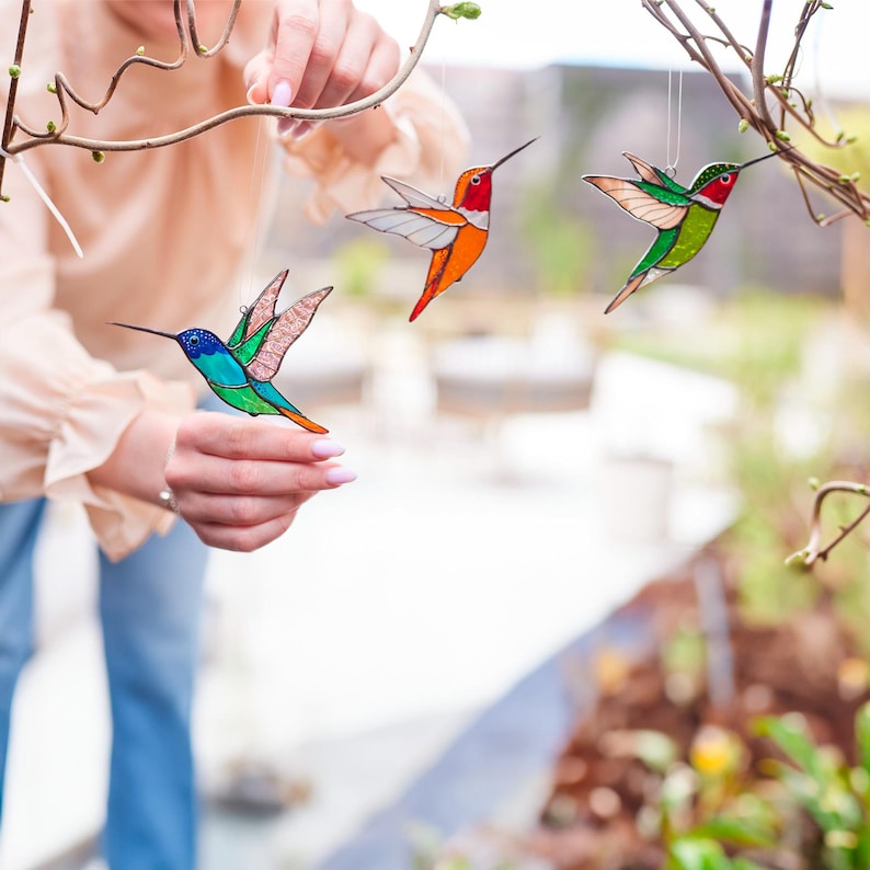 K&ouml;nnte beinhalten: Drei farbenfrohe Glasmalerei-Kolibri-Ornamente, die an einem Ast h&auml;ngen. Die Kolibris weisen lebendige Farbt&ouml;ne von Blau, Gr&uuml;n, Orange und Rot auf. Jeder Vogel hat einen langen Schnabel und ausgebreitete Fl&uuml;gel, was eine dekorative Darstellung ergibt.