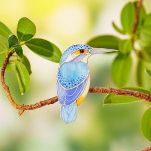 Puede incluir: Un pájaro de vidriera, principalmente azul con acentos amarillos y blancos, posado en una rama marrón. El pájaro tiene un pico largo y está rodeado de hojas verdes, creando un diseño inspirado en la naturaleza.