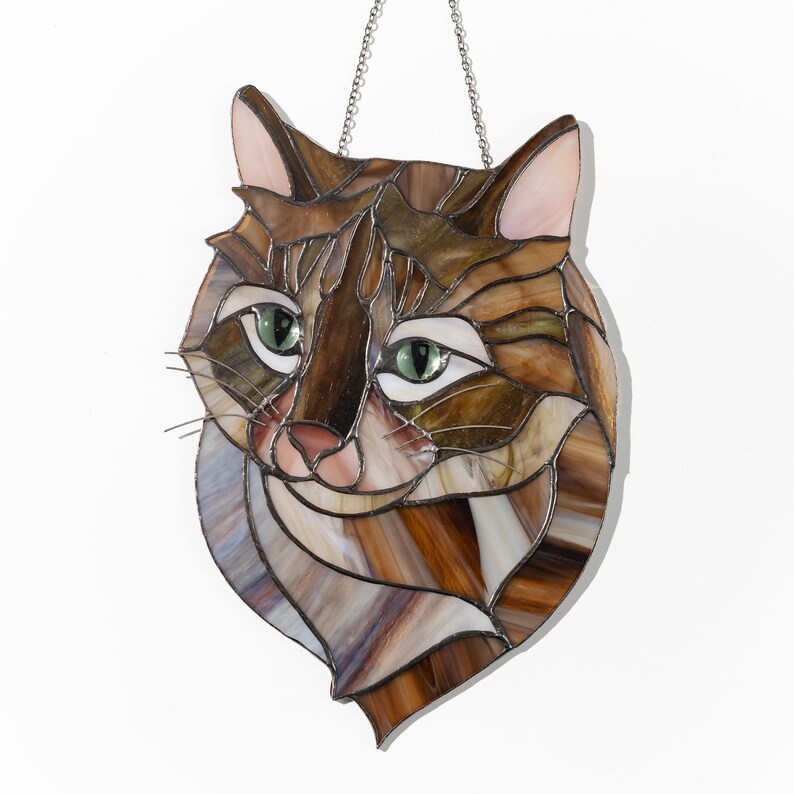 May include: A stained glass window hanging with a brown and white cat face. The cat has green eyes and a pink nose.