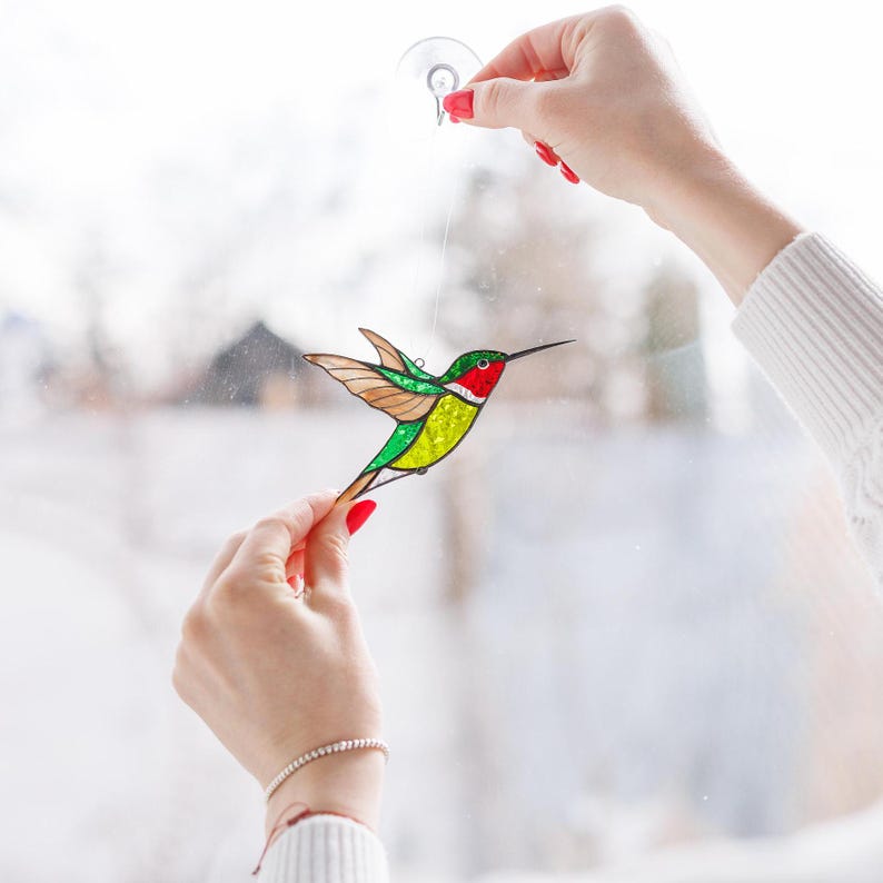 K&ouml;nnte beinhalten: Ein Glasfenster-Kolibri-Sonnenf&auml;nger mit roten, gr&uuml;nen, gelben und braunen Details. Der Kolibri wird von einer durchsichtigen Schnur und einem Saugnapf gehalten. Die Fl&uuml;gel des Vogels sind im Flug ausgebreitet.