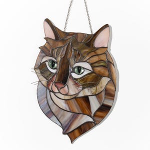 May include: A stained glass window hanging with a brown and white cat face. The cat has green eyes and a pink nose.