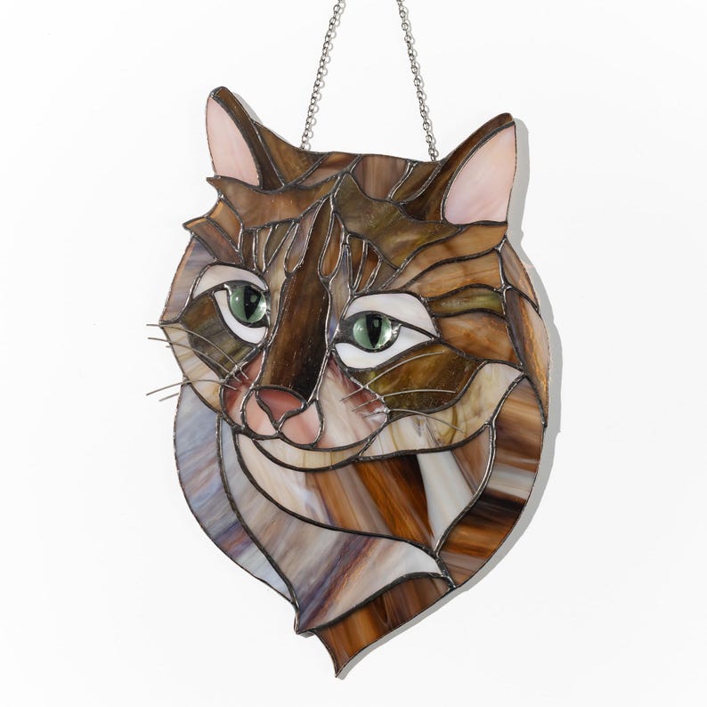 May include: A stained glass window hanging with a brown and white cat face. The cat has green eyes and a pink nose.