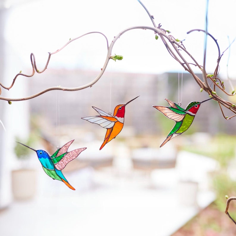 K&ouml;nnte beinhalten: Drei bunte Glasmalerei-Hummel-Ornamente, die an einem Ast h&auml;ngen. Die V&ouml;gel zeigen leuchtende Farben wie Rot, Orange, Gr&uuml;n und Blau. Sie haben lange Schn&auml;bel und ausgebreitete Fl&uuml;gel, was eine dekorative und k&uuml;nstlerische Darstellung ergibt.