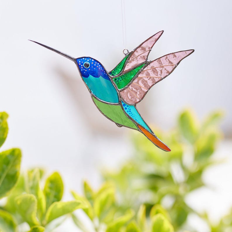 K&ouml;nnte beinhalten: Ein Glas-Hummingbird mit langem Schnabel und ausgebreiteten Fl&uuml;geln. Der Vogel hat einen blauen Kopf mit wei&szlig;en Punkten, einen t&uuml;rkisfarbenen K&ouml;rper, einen gr&uuml;nen Bauch und rosa Fl&uuml;gel. Der Kolibri h&auml;ngt vor einem verschwommenen gr&uuml;nen und wei&szlig;en Hintergrund.