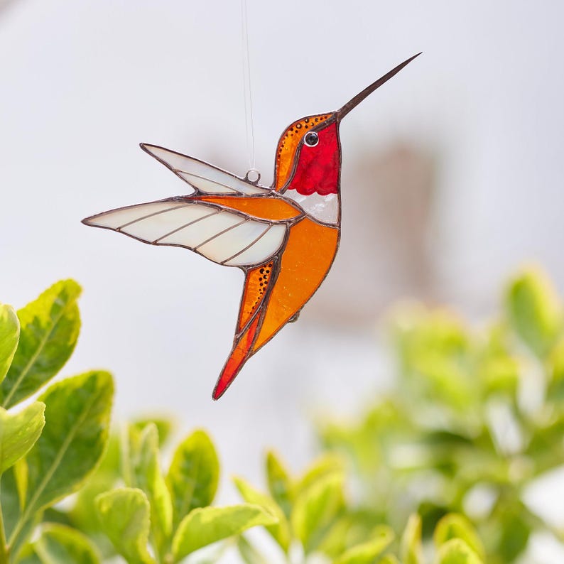 K&ouml;nnte beinhalten: Ein farbenfroher Glas-Hummingbird mit langem Schnabel und ausgebreiteten Fl&uuml;geln. Die Farben sind Rot, Orange und Wei&szlig;. Der Kolibri h&auml;ngt an einem d&uuml;nnen Draht, was darauf hindeutet, dass es sich um einen dekorativen Artikel handelt.
